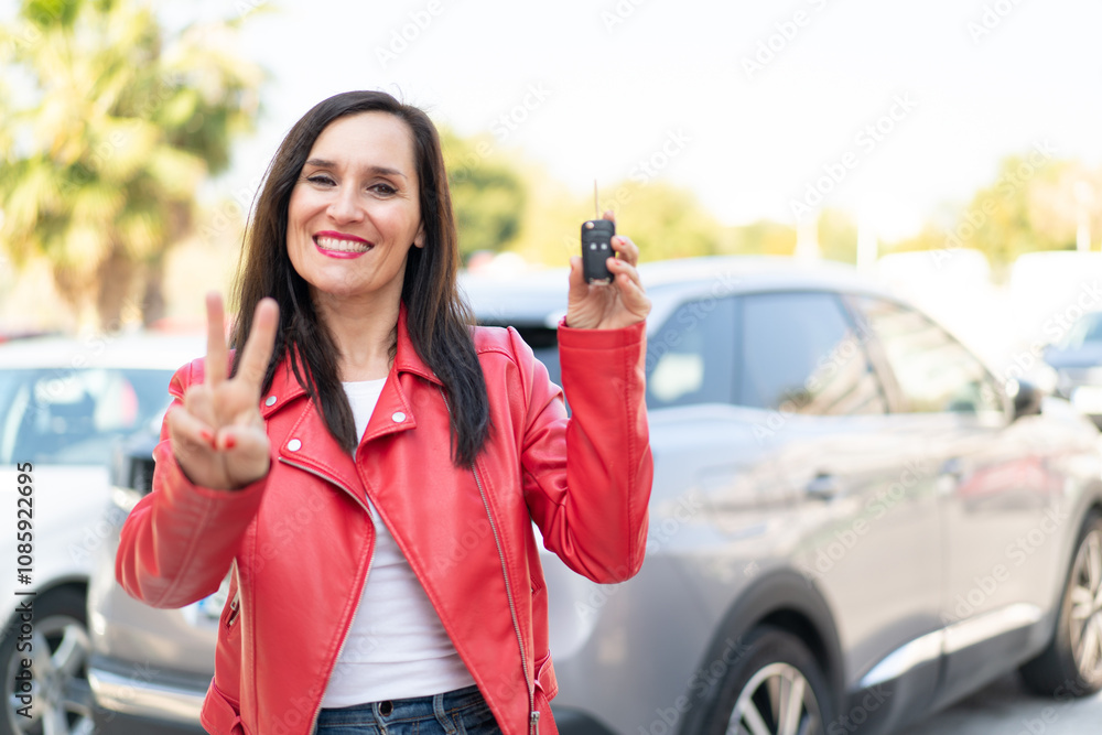 Middle aged woman holding car keys at outdoors smiling and showing victory sign Middle aged woman holding car keys at outdoors smiling and showing victory sign