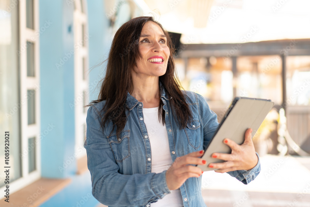 Middle aged woman holding a tablet at outdoors looking up while smiling Middle aged woman holding a tablet at outdoors looking up while smiling