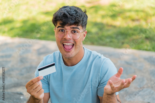 Young hispanic man holding a credit card at outdoors with shocked facial expression photo