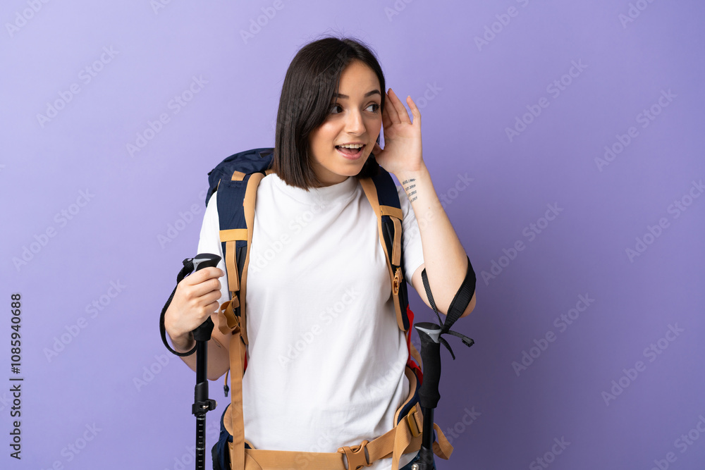 Young caucasian woman with backpack and trekking poles isolated on blue background listening to something by putting hand on the ear Young caucasian woman with backpack and trekking poles isolated on blue background listening to something by putting hand on the ear