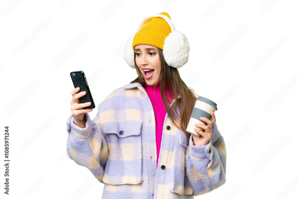Young woman wearing winter muffs over isolated chroma key background holding coffee to take away and a mobile Young woman wearing winter muffs over isolated chroma key background holding coffee to take away and a mobile