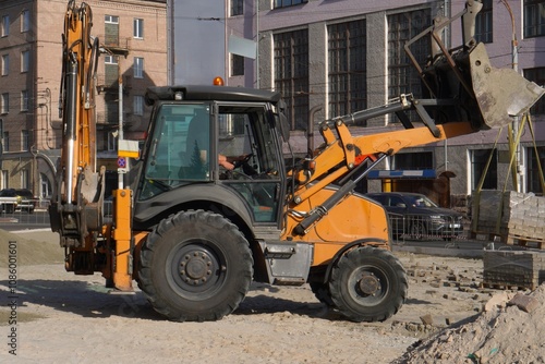 A yellow tractor repairs a road in the city with a bucket and tiles near the roadway during the day