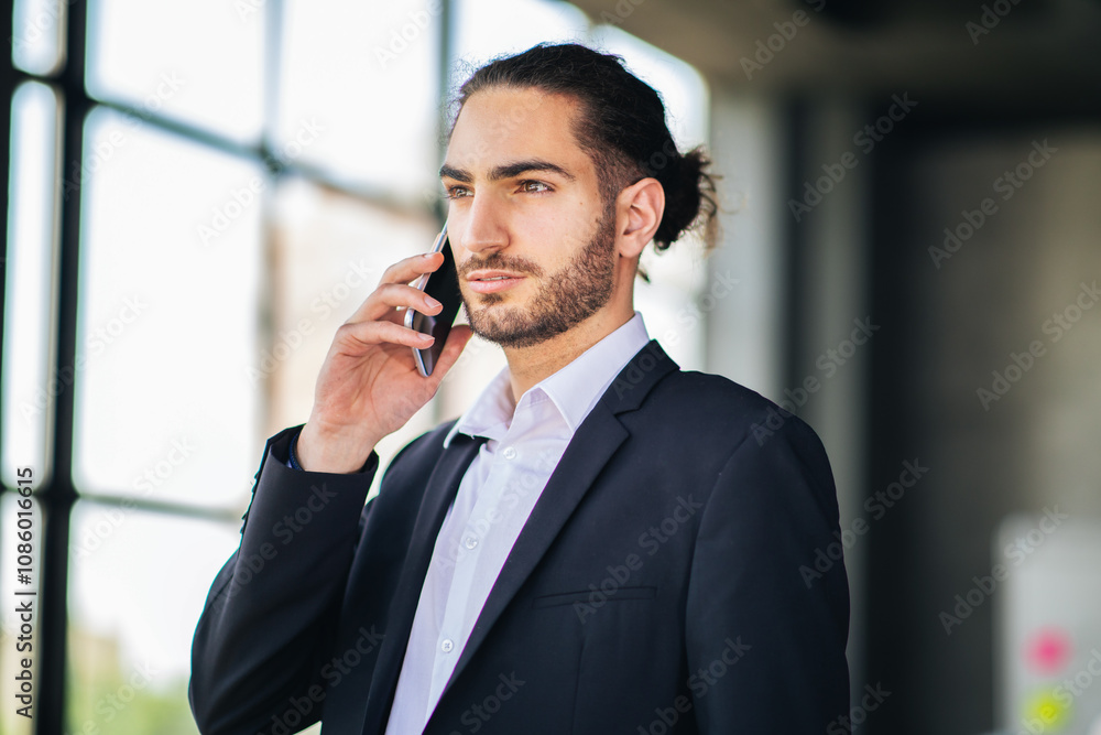 A business professional wearing a suit is talking on a mobile phone inside a modern office setting. Sunlight streams through large windows, creating a dynamic atmosphere. A business professional wearing a suit is talking on a mobile phone inside a modern office setting. Sunlight streams through large windows, creating a dynamic atmosphere.