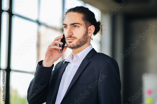 A business professional wearing a suit is talking on a mobile phone inside a modern office setting. Sunlight streams through large windows, creating a dynamic atmosphere.