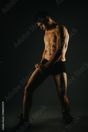 Fit male posing in a dark studio, highlighting his toned abs, muscular arms, and overall fit body.