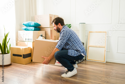 Quadro em tela Profile view of a man squatting and lifting a heavy cardboard box with care as h