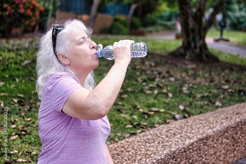 Gray-haired elderly lady drinks water from a bottle in the park.