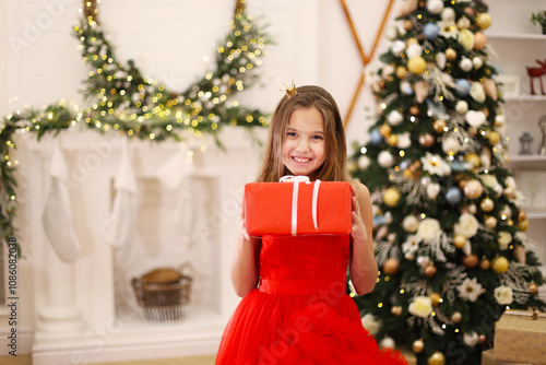 Smiling Girl with Gift by Christmas Tree. A cheerful girl in a red dress holds a wrapped gift in a festive room decorated with a Christmas tree and glowing lights. photo