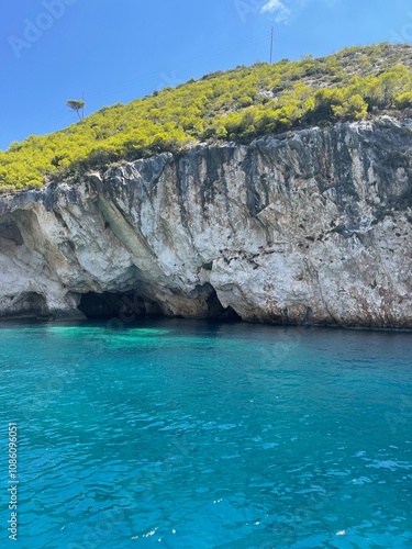 Crystal-clear blue waters of Zakynthos with a majestic cliff rising in the background, showcasing the island’s stunning natural beauty and dramatic landscapes.