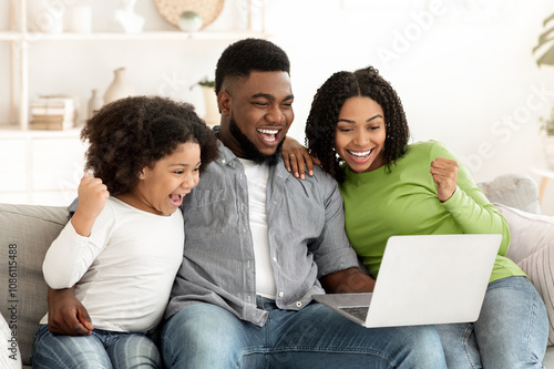 Online Lottery. Excited African American Family Of Three Celebrating Win With Laptop, Sitting On Couch In Modern Living Room