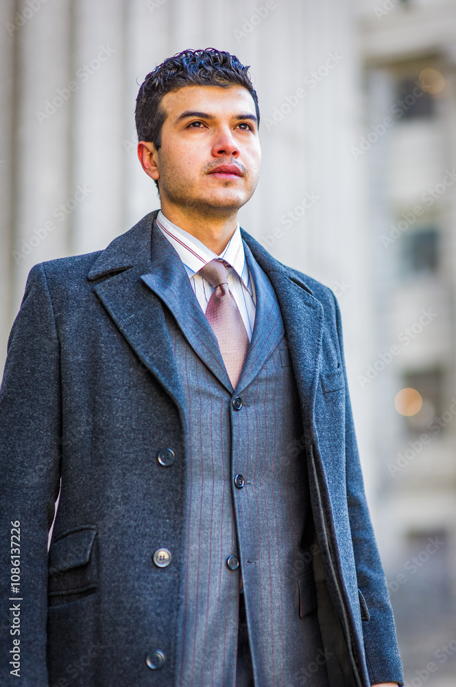 A man in a stylish gray coat and suit stands confidently outdoors, looking into the distance. The background features blurred architectural elements that suggest an urban setting..
