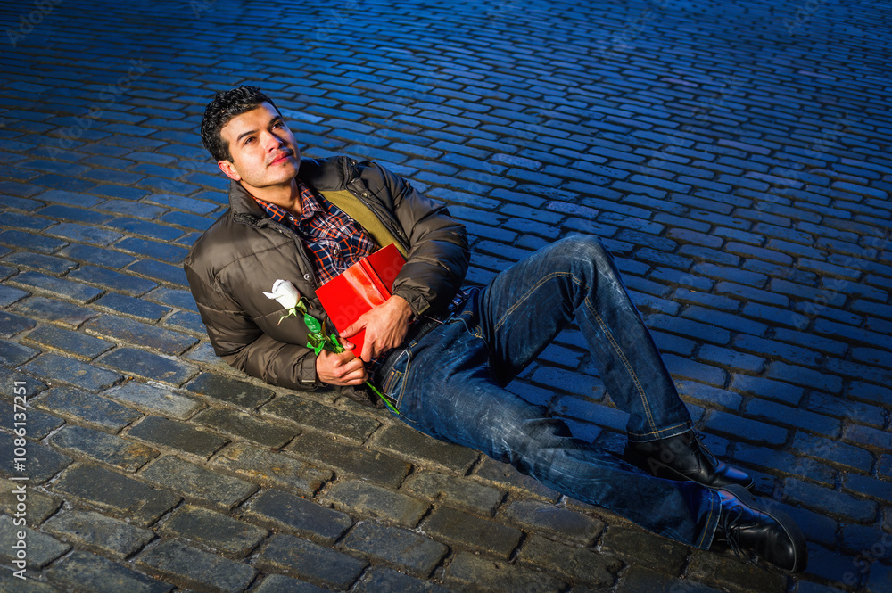 A man is lying on a cobblestone street, looking contemplative while holding a red book and a white rose. He is dressed in a casual jacket and jeans, and is illuminated by dramatic lighting.