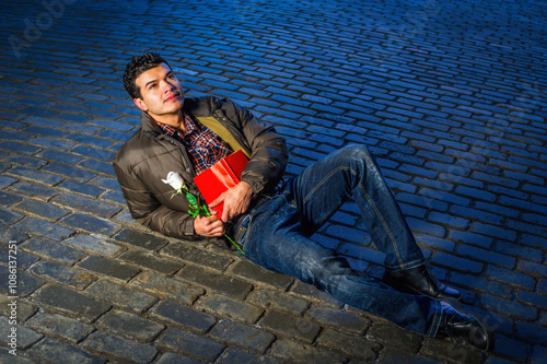 Obraz na płótnie A man is lying on a cobblestone street, looking contemplative while holding a red book and a white rose. He is dressed in a casual jacket and jeans, and is illuminated by dramatic lighting. w salonie