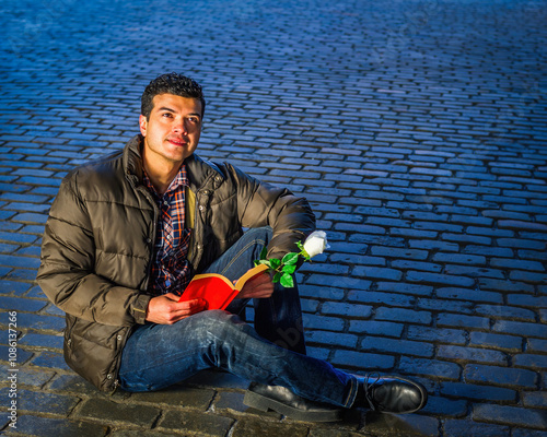 Obraz na płótnie A man in a casual jacket sits on a cobblestone path, holding a white rose and an open book. He gazes thoughtfully into the distance, illuminated by soft, cool lighting.. w salonie