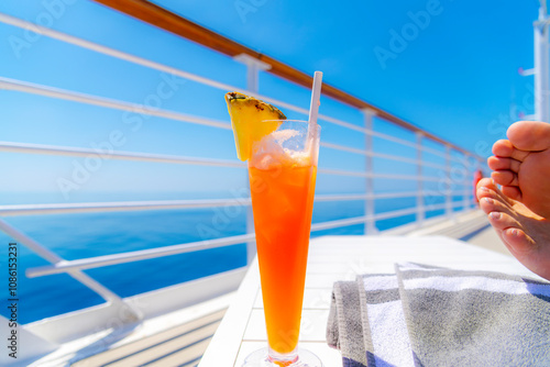 A female cruise ship passenger relaxes with her feet up and a tropical cocktail drink with fresh pineapple, on the upper deck of a large cruise ship at sea.