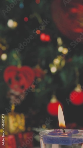Close up of burning candle in focus with blurred decorated Christmas tree with blinking lights. Vertical video, details. Scene background.