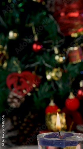 Close up of burning candle in focus with blurred decorated Christmas tree with blinking lights. Vertical video, details. Scene background.