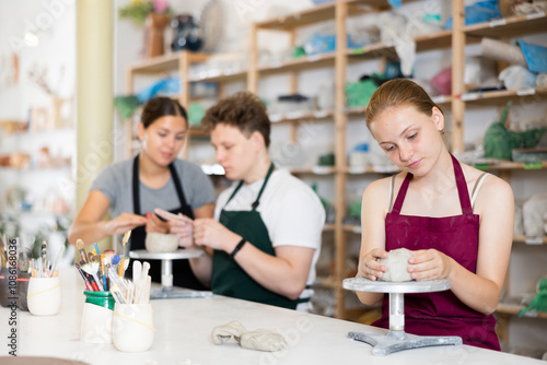 Young female teacher in apron helps teenager boy and teenager girl students to make product from clay in ceramic workshop