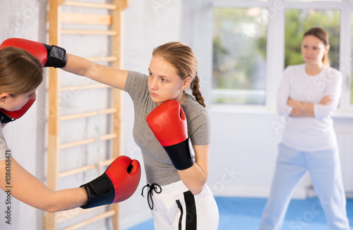 Female partner and girl in boxing gloves strike deliver hook each other, opponent during training bout, improving fighting skills during boxing training. Woman coach watches and comments on battle photo