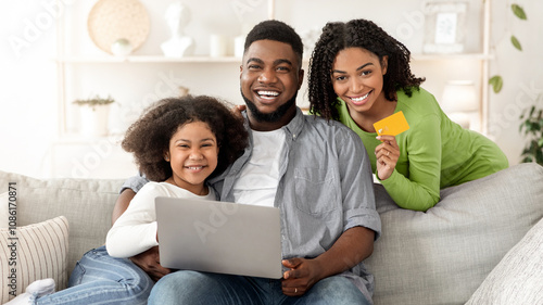 Online Shopping. Happy Black Family Posing With Laptop And Credit Card At Home, Purchasing Goods In Internet Together