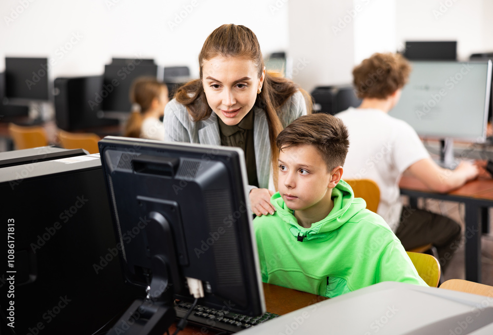 Positive female teacher helping focused preteen schoolboy studying in college library computer lab.. Positive female teacher helping focused preteen schoolboy studying in college library computer lab..