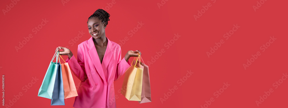 Fototapeta premium Happy young African American woman holding colorful shopping bags in her hands, posing in pink costume on red background, panorama with copy space