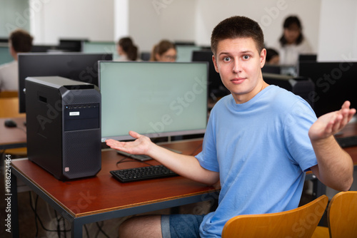 Thoughtful teenage boy sitting at desk in computer class and making helpless gesture. Young man studying computer science. photo