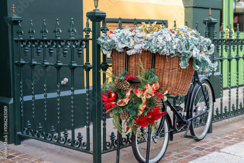 A black two-wheel bicycle chained to a dark green wrought iron fence. The butcher's bike has a large wicker basket filled with greenery and Christmas flowers. There's a wreath with red bows on front.  photo