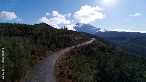 A scenic aerial view of a winding road surrounded by lush forested hills leading toward a majestic Mount Rainier under a blue sky with scattered clouds, capturing the beauty of the Pacific Northwest.