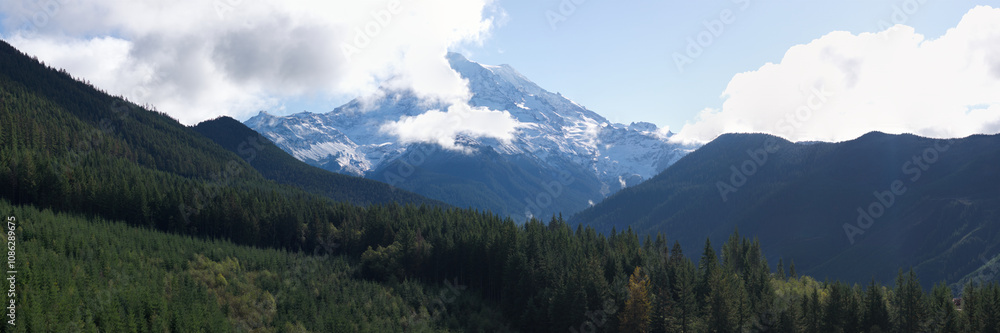 Obraz premium A scenic panorama capturing Mount Rainier partially covered by clouds, surrounded by lush, forested hills under a bright sky.