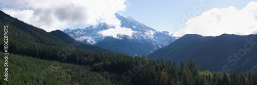 A scenic panorama capturing Mount Rainier partially covered by clouds, surrounded by lush, forested hills under a bright sky.