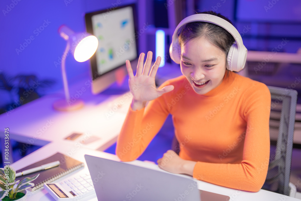 Smiling young woman wearing headphones waves at her laptop during a ...