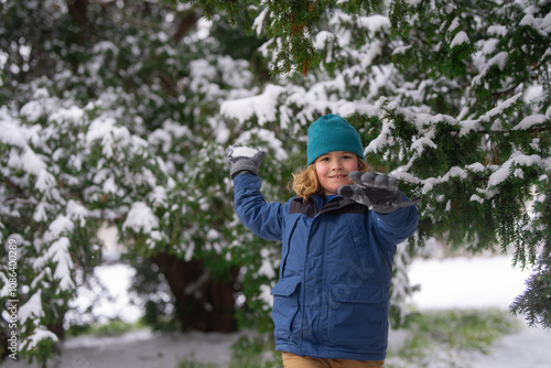 Child play with snow in winter park. Kids with first snow. Child winter holiday. Smiling boy playing with snow in winter park. Portrait of a little kid in knit hat throw snowballs. Funny kids in snow.