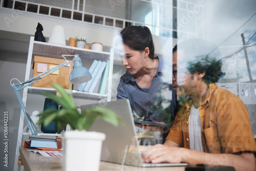 Fotografie View from outside reflecting window, two young Asian startup company partners discuss and brainstorm in a casual small business office, marketing planning jobs on a laptop, and SME freelance teamwork