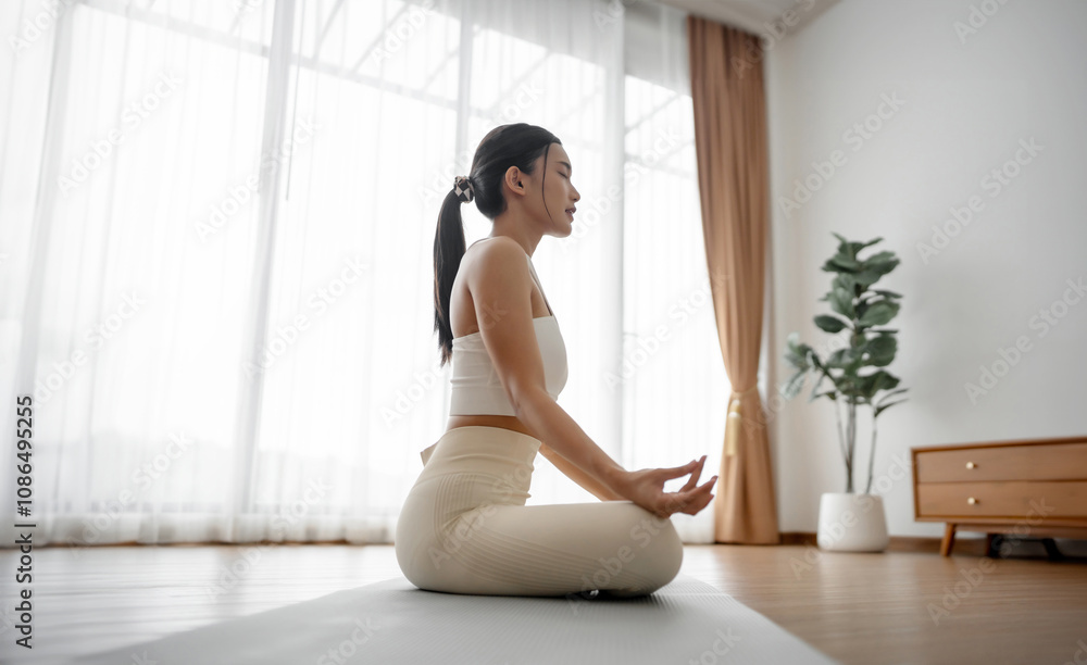 Young Asian woman meditating in lotus position at home, sitting on the floor in fitness clothing. Breathing workout to achieve relaxation, peace and mindfulness Young Asian woman meditating in lotus position at home, sitting on the floor in fitness clothing. Breathing workout to achieve relaxation, peace and mindfulness
