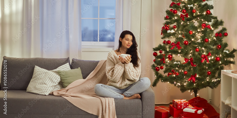 A woman enjoys a warm drink while seated comfortably on a sofa next to a lit Christmas tree. This image captures the cozy and festive spirit, ideal for holiday-themed marketing or relaxation content. A woman enjoys a warm drink while seated comfortably on a sofa next to a lit Christmas tree. This image captures the cozy and festive spirit, ideal for holiday-themed marketing or relaxation content.