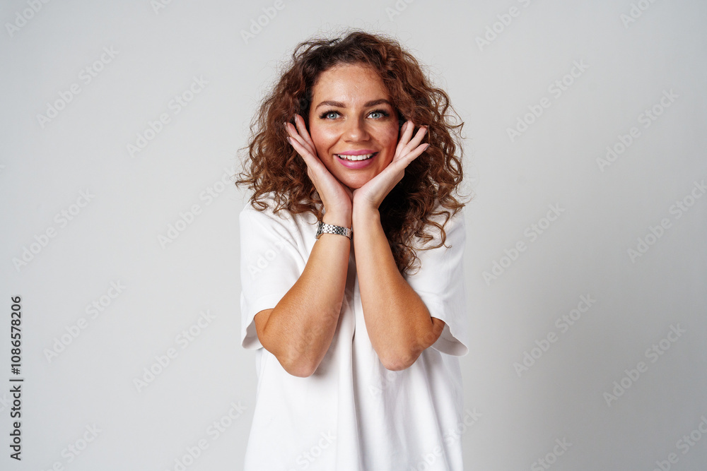 Woman with curly hair smiles joyfully while posing with hands on her cheeks against a light background Woman with curly hair smiles joyfully while posing with hands on her cheeks against a light background