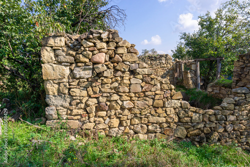 An old ruined wall made of large stones illuminated by sunlight. Grass and trees around