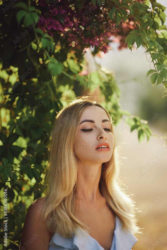 Fototapeta premium Beautiful blonde woman with closed eyes enjoying sunlight, surrounded by lush green leaves and purple flowers. Perfect for concepts of relaxation, beauty, and nature, capturing a serene moment.