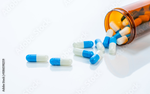 Prescription antibiotic capsules spilling from amber bottle onto a white background. Blue and white capsule pills. Ideal for healthcare, pharmaceutical treatments, and medical medication industries.