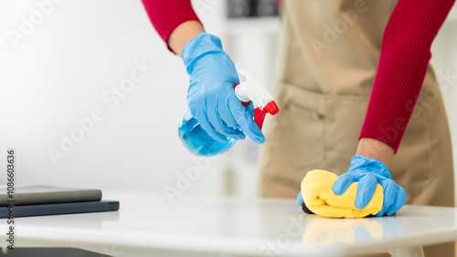 A young woman cleans her desk at home, using a disinfectant spray and a wipe. She ensures a hygienic and organized workspace, emphasizing cleanliness and personal health in her routine