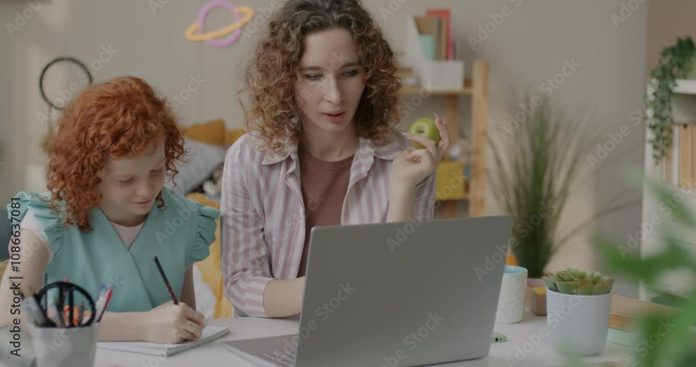 Caring mother teaching cute little daughter looking at laptop screen and writing in notebook at home. Family and education concept. Caring mother teaching cute little daughter looking at laptop screen and writing in notebook at home. Family and education concept.