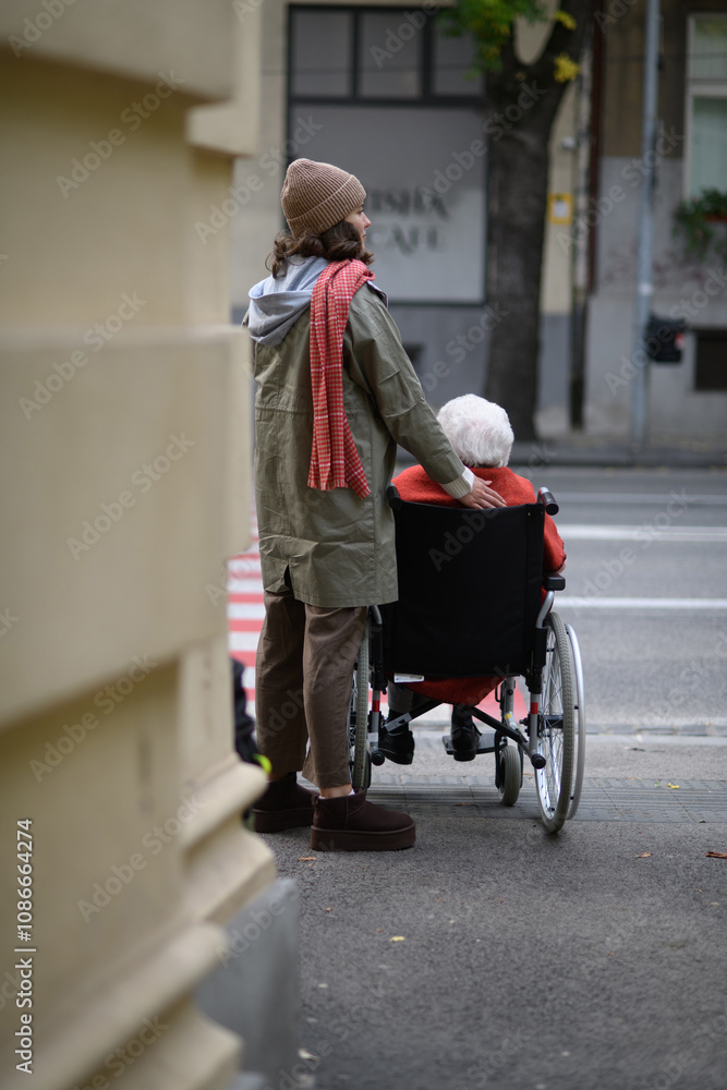 Obraz premium Granddaughter on an autumn walk in the city with her grandmother, pushing her in wheelchair. Rear view.