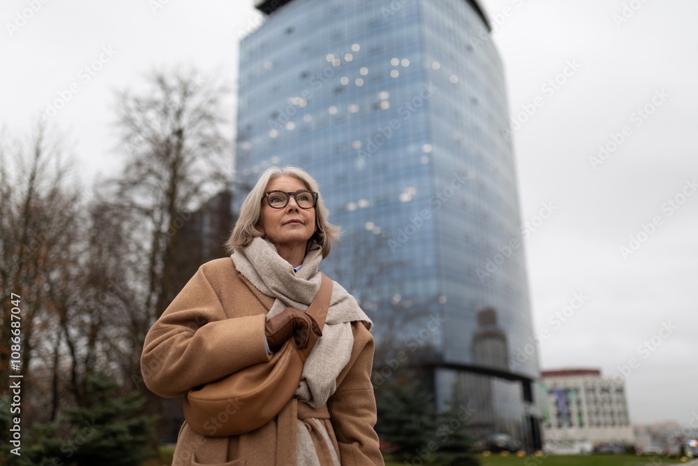 A woman in a beige coat walks thoughtfully past a modern glass building on a cloudy day in an urban setting A woman in a beige coat walks thoughtfully past a modern glass building on a cloudy day in an urban setting