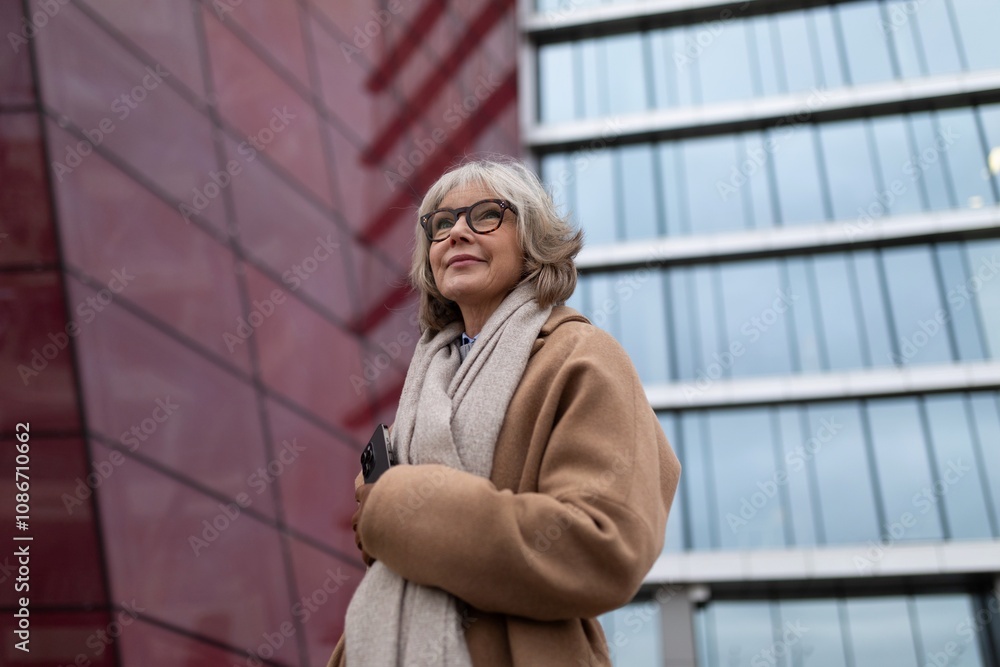 A smiling older woman in a stylish coat stands outside a modern glass building on a cloudy day, showcasing confidence and elegance A smiling older woman in a stylish coat stands outside a modern glass building on a cloudy day, showcasing confidence and elegance