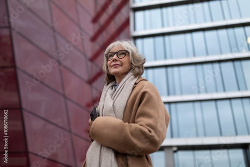 A smiling older woman in a stylish coat stands outside a modern glass building on a cloudy day, showcasing confidence and elegance