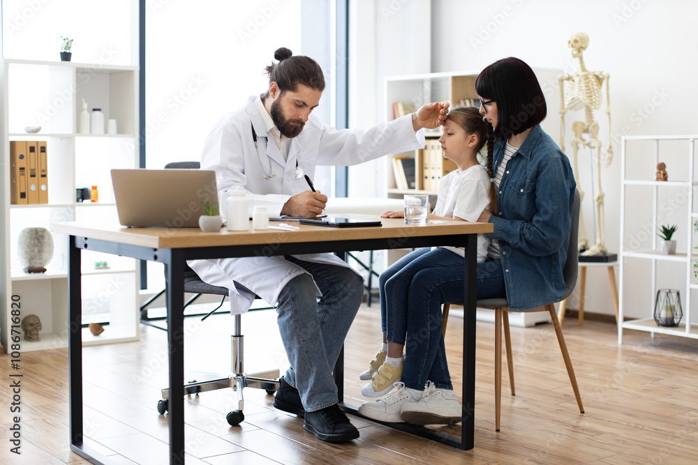 Caucasian male doctor measures temperature by touching forehead of young girl in clinic. Female caregiver sits by girl's side while doctor writes prescription. Caucasian male doctor measures temperature by touching forehead of young girl in clinic. Female caregiver sits by girl's side while doctor writes prescription.