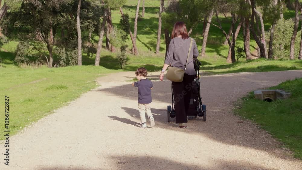 Woman walking in nature on a sunny day with a baby stroller and a small child