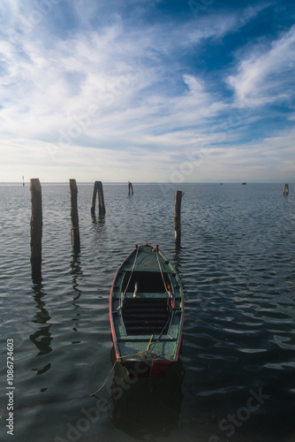 Una piccola barca di legno dipinta di verde ormeggiata a un palo davanti a Pellestrina, isola della laguna di Venezia, in una giornata autunnale con cielo azzurro e nuvole