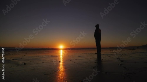 silhouetted man watching sun set into ocean standing on reflective wet sand camera tracking 